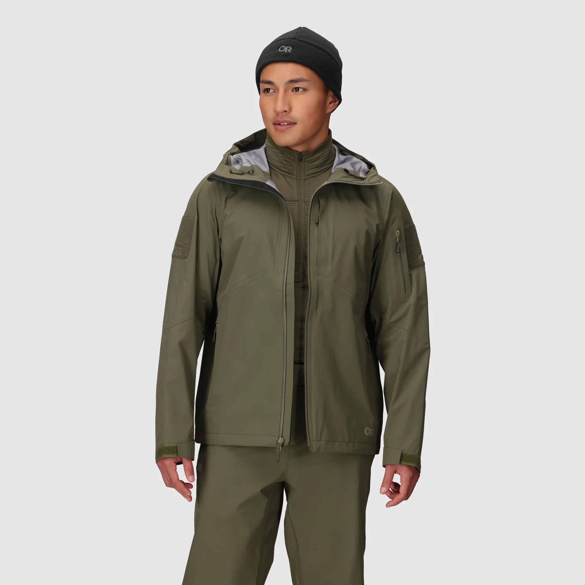 Person wearing a green raincoat and pants with a black beanie on a light gray background