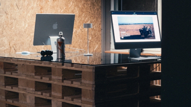 Two computer monitors on a wooden desk with a rustic wall background